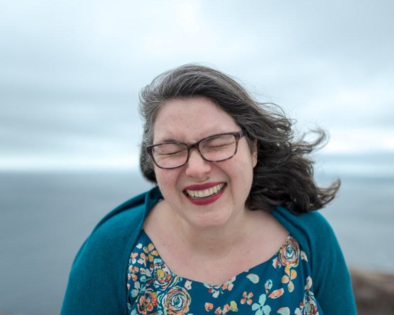 Image: Andreae, a white woman with shoulder-length salt-and-pepper hair, laughs into the camera. Her hair and clothes are whipped by the wind, and her eyes are squeezed shut behind her glasses. She wears a teal floral dress and teal sweater. In the background, the ocean and sky are slate grey. Photo credit: m+e photography