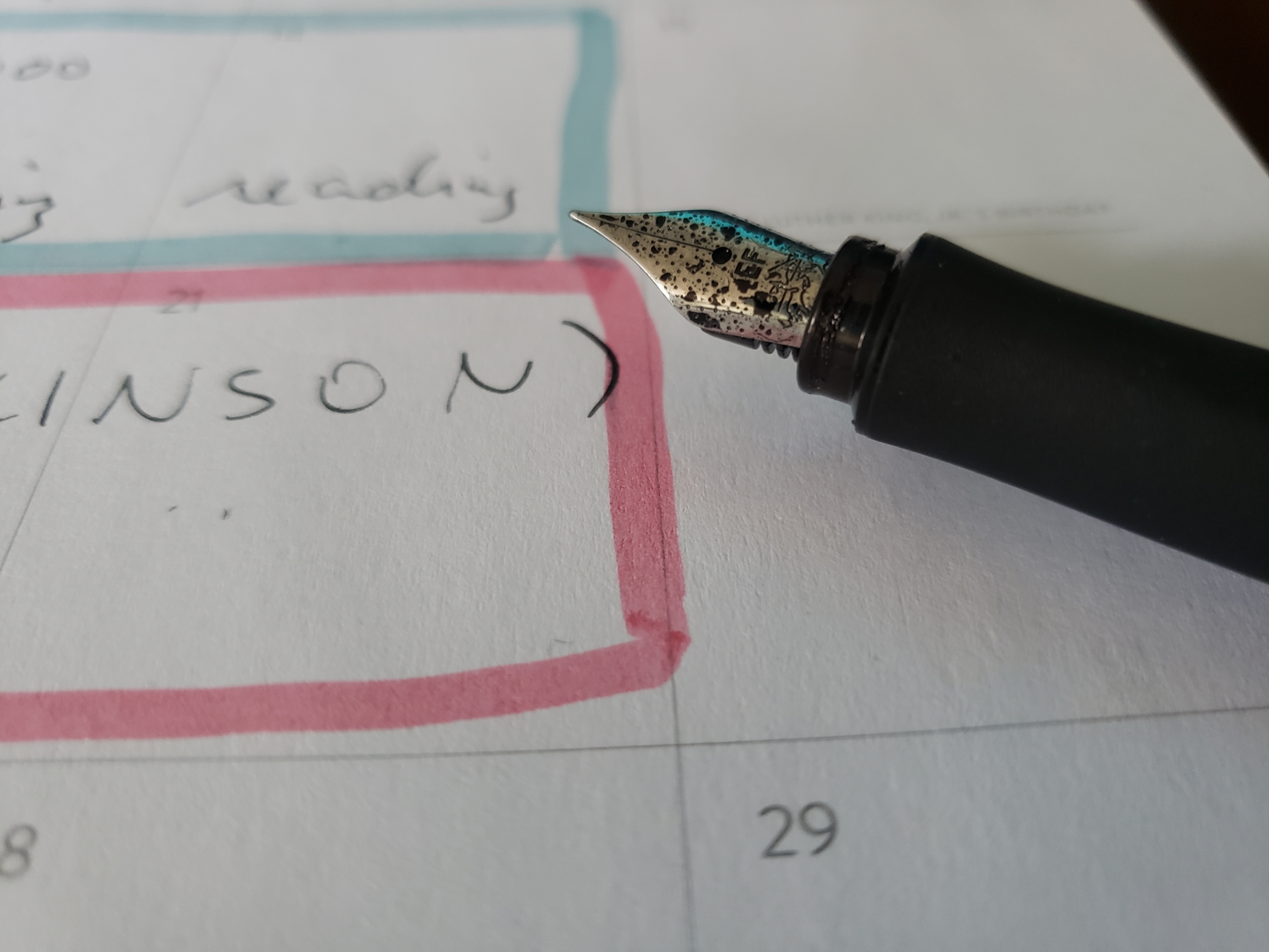 A close-up of a fountain pen resting on the the page of a planner open to a calendar spread. The nib of the pen is dotted with ink splatters. The planner page has days outlined in muted blue and pink highlighter, and the tasks written in the squares are scrawled in messy handwriting.