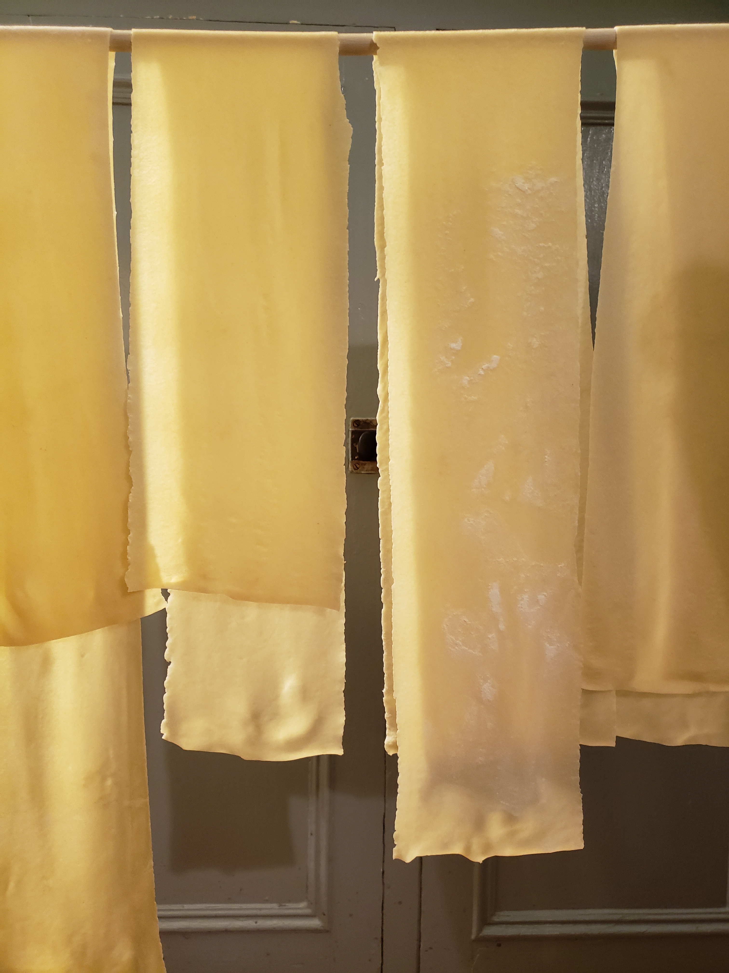 Four long, wide strips of fresh pasta are draped over a horizontal dowel. Behind them is a blue-grey wooden cabinet. 