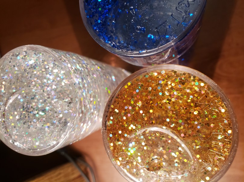 A view from above of three sensory glitter tubes standing upright on a desk. The tubes are made of clear plastic and are about the same dimensions as those skinny soft drink cans you can get now. They are filled with clear liquid and coloured glitter. The one in the lower right is filled with gold glitter; the one to the left, silver glitter; the one in the upper right, royal blue glitter. 