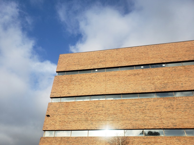 The top storeys of a brick building -- Memorial University's QEII library -- shot from below. The building is striped with horizontal lines of reflective window glass. The yellow-brown bricks stand out against a blue and white winter sky. In the lower windows, treetops are reflected; in the upper ones, clouds. 