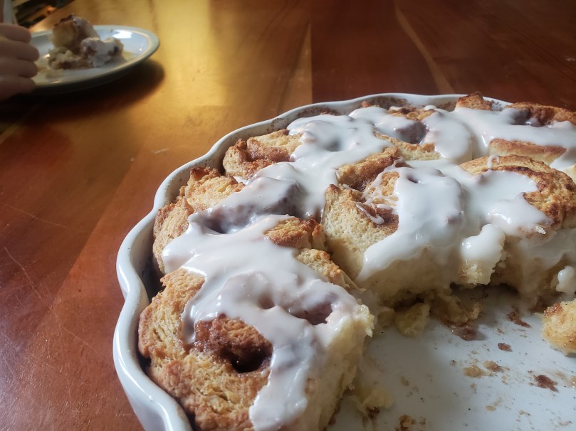 A batch of delicious-looking, buttery, flaky cinnamon buns in a white scalloped quiche pan. They are frosted with a white icing-sugar glaze. Several of the cinnamon buns have already been removed from the pan. In the upper left of the photo, a half-eaten cinnamon bun sits on a plate; a child's hand holding the plate still suggests that the remainder of the bun will soon be eaten as well.