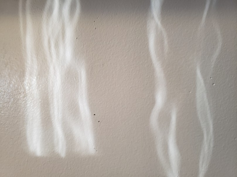 Two sets of wavy vertical light streaks on a grey-painted and slightly pitted interior wall. The light is wavy from having passed through the panes of an old French door.