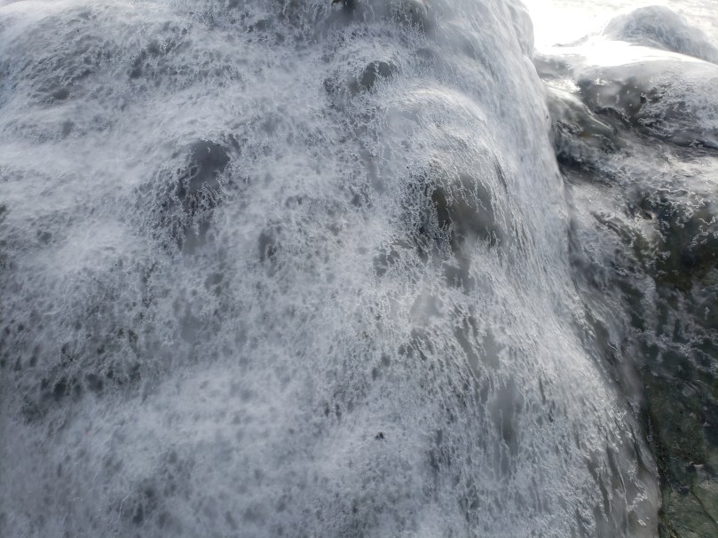 A large boulder on a beach is encrusted with a layer of clear ice. The ice is veined with white and looks lace-like against the grey-black of the stone.