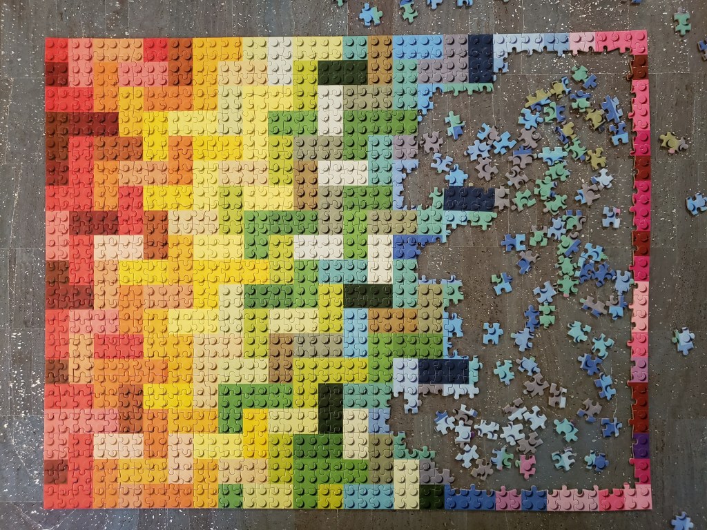 View from above of a mostly completed LEGO themed jigsaw puzzle. The puzzle image is of 2x2 and 2x4 LEGO pieces arranged in a quilt-like pattern. The colour scheme of the puzzle moves from orange-reds on the left, through yellows, greens, blues, purples and, at the far right, pinks. The puzzle is laid out on a greyish cork surface flecked with silver. 
