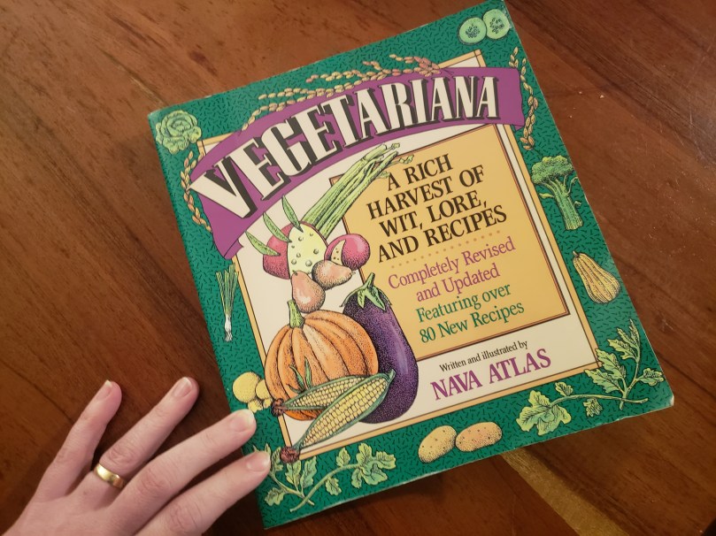 A photograph of a cookbook resting on a wooden tabletop. A hand -- white skin, short unpainted nails, plain gold wedding band -- is touching the bottom left corner of the book. The book cover reads, "Vegetariana: A Rich Harvest of Wit, Lore, and Recipes. Completely Revised and Updated, Featuring over 80 New Recipes. Written and Illustrated by Nava Atlas." The book is square-shaped, and the colours green, gold, and purple dominate.