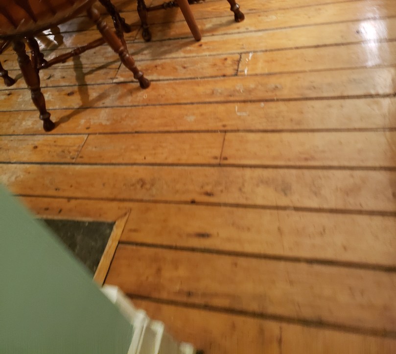 A slightly out-of-focus, weirdly angled shot of a softwood floor. The legs of three wooden country-style dining chairs are visible at the upper left corner.