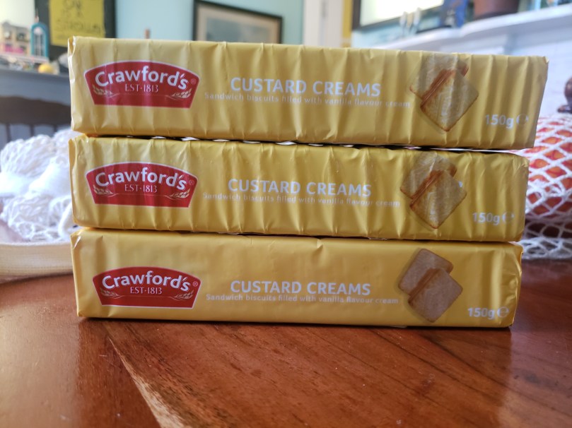 A stack of three 150 gram sleeves of Crawford's Custard Creams. Their packaging is an appropriately custardy yellow; at the right end of each sleeve is a red logo that says "Crawford's EST 1813," and on the left end is a photography of two rectangular sandwich cookies. The three packets are sitting on a dining room table; just behind them are several cotton net shopping bags. Beyond them, the general clutter of daily life.