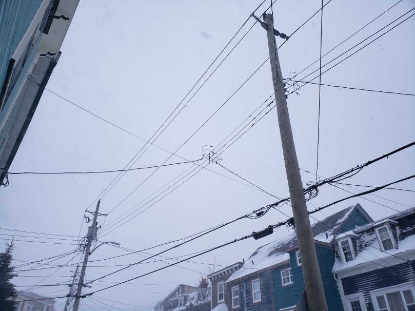 Looking up at poles, wires, rooftops, and a grey sky. It's hard to tell in the photo, but there is snow falling toward the camera. The houses are dusted with snow as well. 