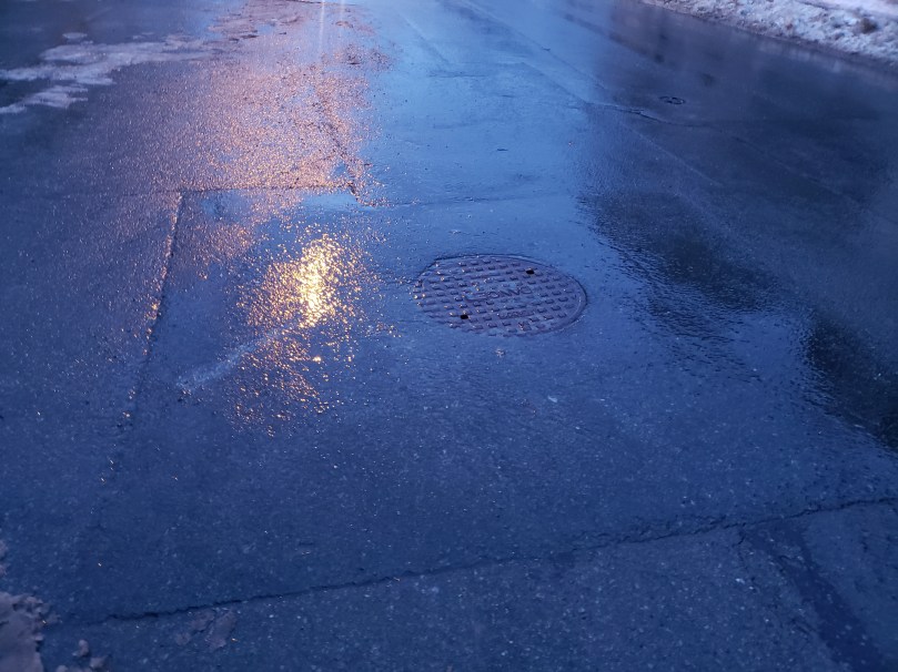 Yellow-orange light from a streetlamp reflects on wet asphalt. The asphalt is patchy from years of summer repairs, and there is a circular manhole cover in the middle of it (covering a manhole, not just sitting there). It is twilight and the road looks bluey-purple. There are traces of dirty snow visible at the upper corners of the photo.