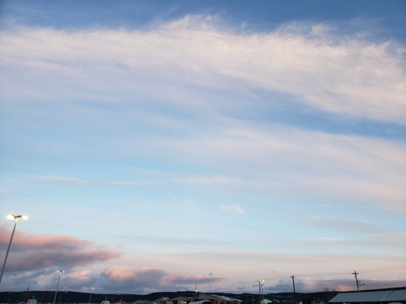 Early evening sky over the Dominion parking lot. The sky is a medium blue at the top of the photo, fading to a watery, almost yellowy grey near the bottom. Most of the sky is streaked with high, thin white clouds, and there is a bank of low cumulus clouds closer to the horizon -- these are purplish and lit with coral-orange light from the setting sun. Streetlights and the tops of houses are visible along the very bottom edge of the photo.