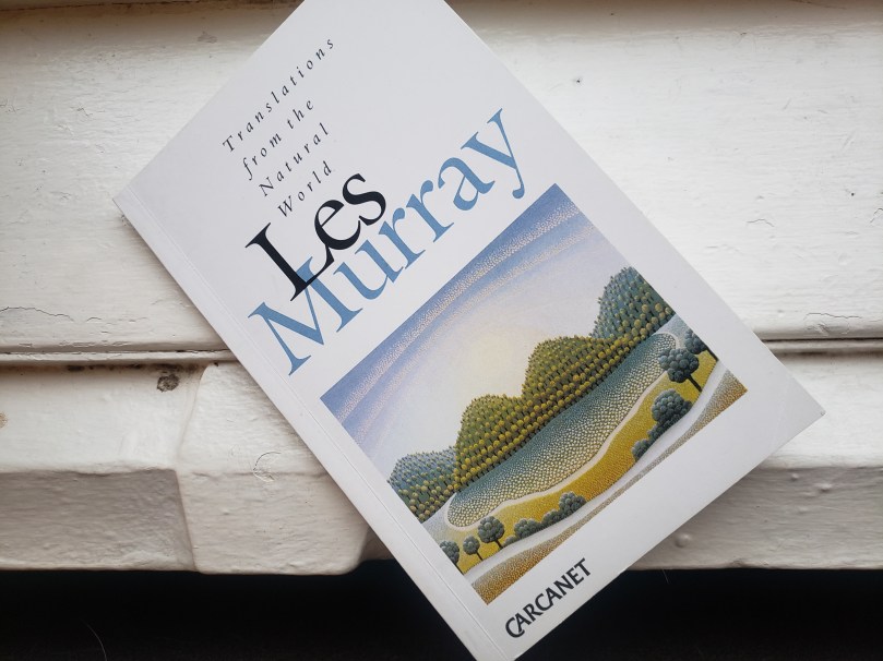 A book -- Translations from the Natural World by Les Murray -- sits on a white-painted windowsill. The book cover is white, with the title in fairly small italicized letters and the author name in much larger type. On the lower half of the book cover is a painting by Croatian artist Ivan Rabuzin. The painting is rendered in the Naïve style, and depicts four tree-covered hills under a bright sky with arching clouds.  
