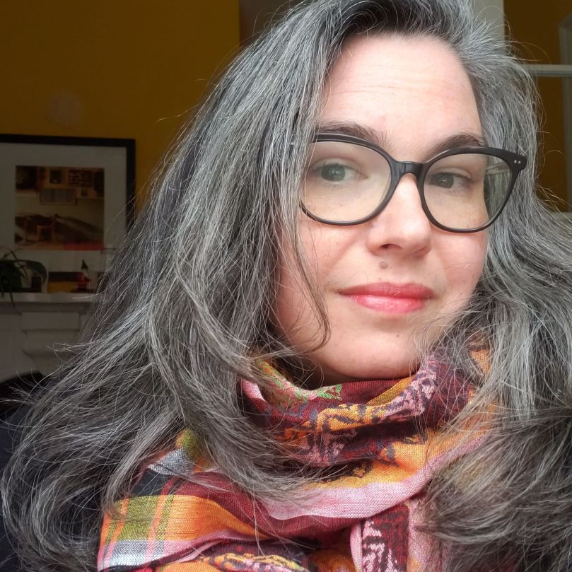 Andreae, a white woman with glasses and long, salt-and-pepper hair, is looking at the camera. She is wearing a pink, burgundy, and orange striped kufiya with green and grey details. 