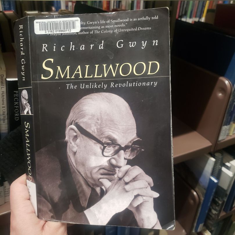 Andreae's hand holds up a paperback copy of Richard Gwyn' s book, Smallwood: The Unlikely Revolutionary. From the background, you can tell that this is occurring in the university library stacks. The book cover is in black and white, and it has a photo of an aging Joey Smallwood -- a mostly bald white man with thick-framed glasses and unruly eyebrows -- resting his chin on his thumbs while his hands are folded in front of him. He is looking pensive.