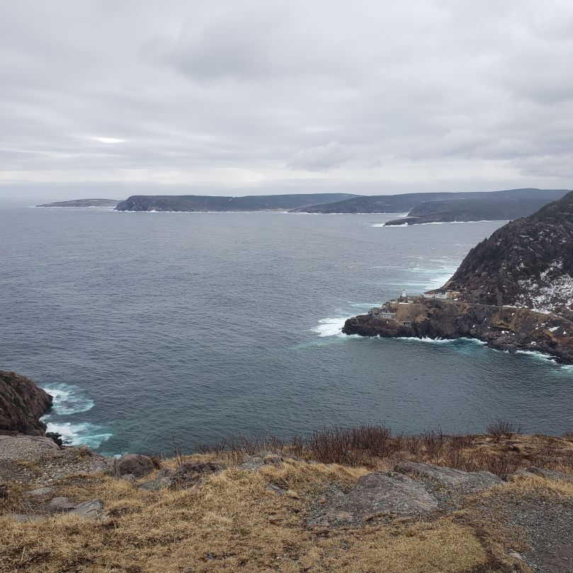 A bleak view of the Atlantic Ocean, looking from Signal Hill past Fort Amherst. The grass is yellowed and scrappy, and patches of old snow cling to the rocks above the Fort Amherst lighthouse. The ocean is disturbed, but not choppy. The sky is grey.
