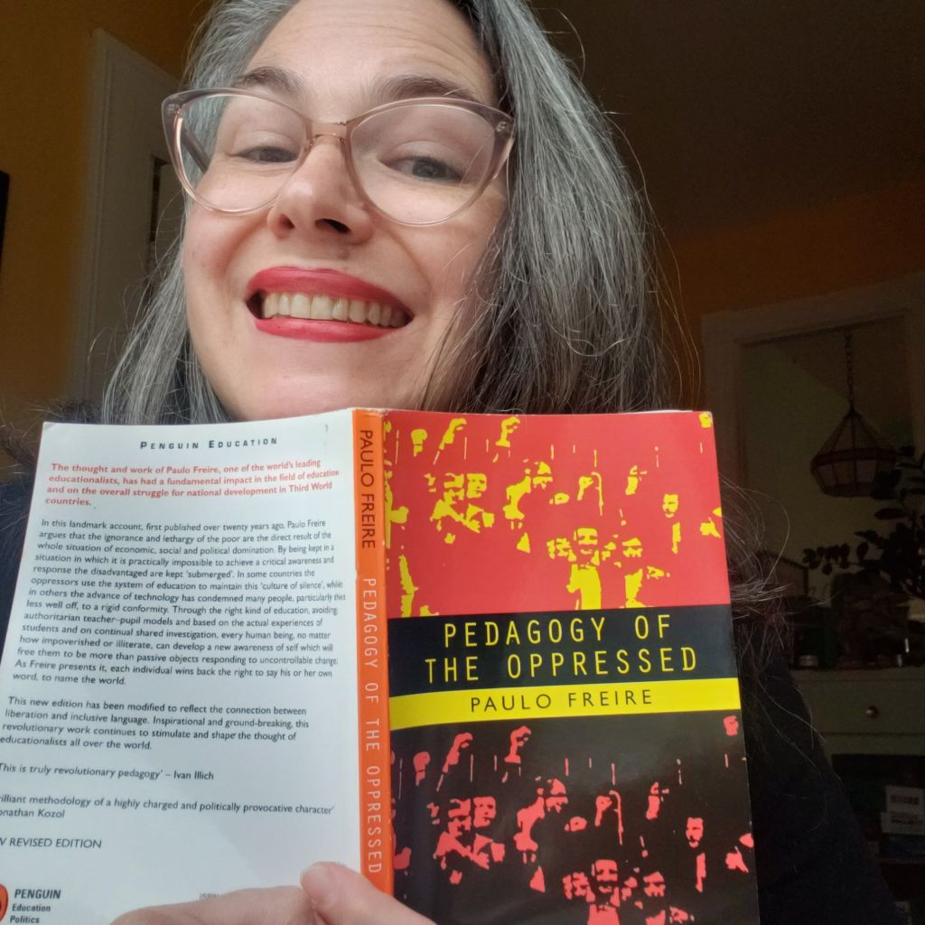 Andreae is smiling while holding up an open copy of the Penguin edition of Pedagogy of the Oppressed by Paula Freiere. The cover of the book is red, yellow, and black, and the spine is orange.
