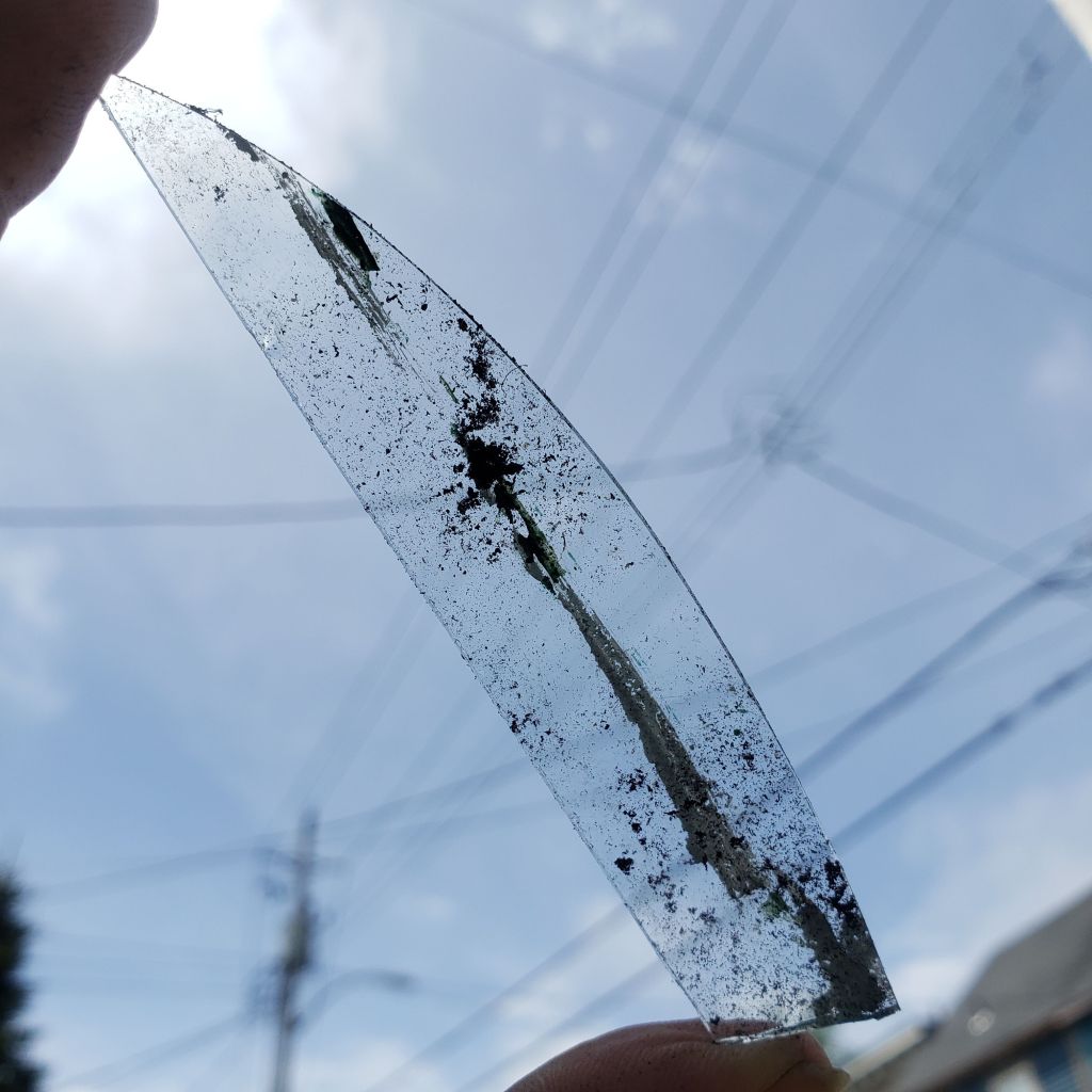 A shard of broken window glass is held against the sky. The glass is about three inches long and three-quarters of an inch wide. There is a streak of dirt running vertically along the glass. The sky in the background is crisscrossed with power lines.  