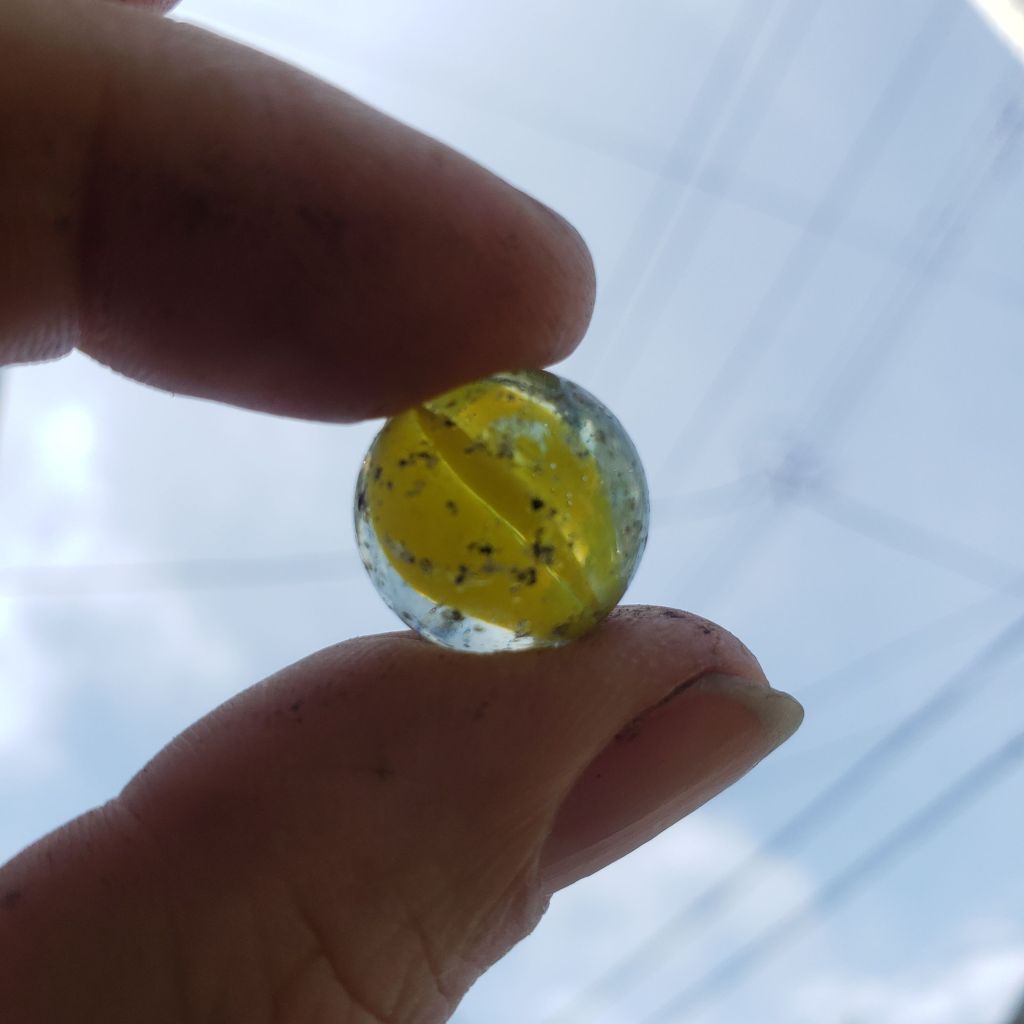 A yellow cat's-eye marble is held between Andreae's thumb and index finger. The marble is against a blue sky crisscrossed with power lines. The surface of the marble is pitted with age.
