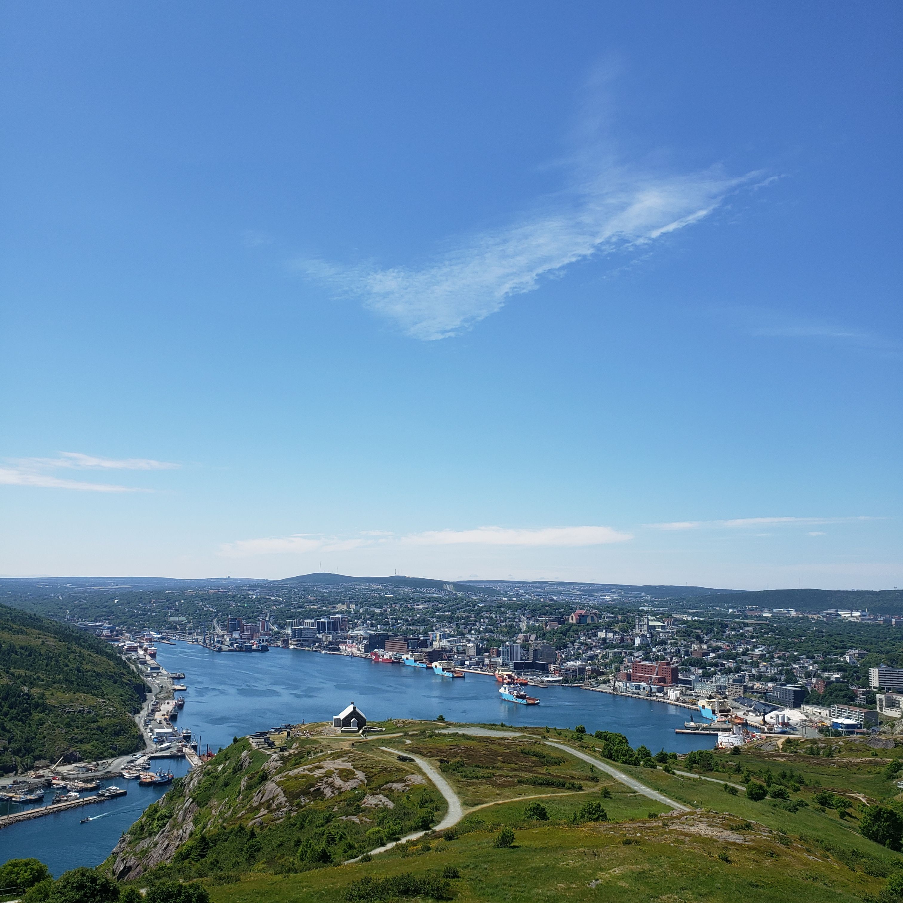 A view of St. John's Harbour from atop Signal Hill. The water is bright blue. The sky is also bright blue, and streaked with the thinnest white clouds. The harbour is flanked by ships on both sides. In the foreground there is green grass cut through by well-maintained trails. 
