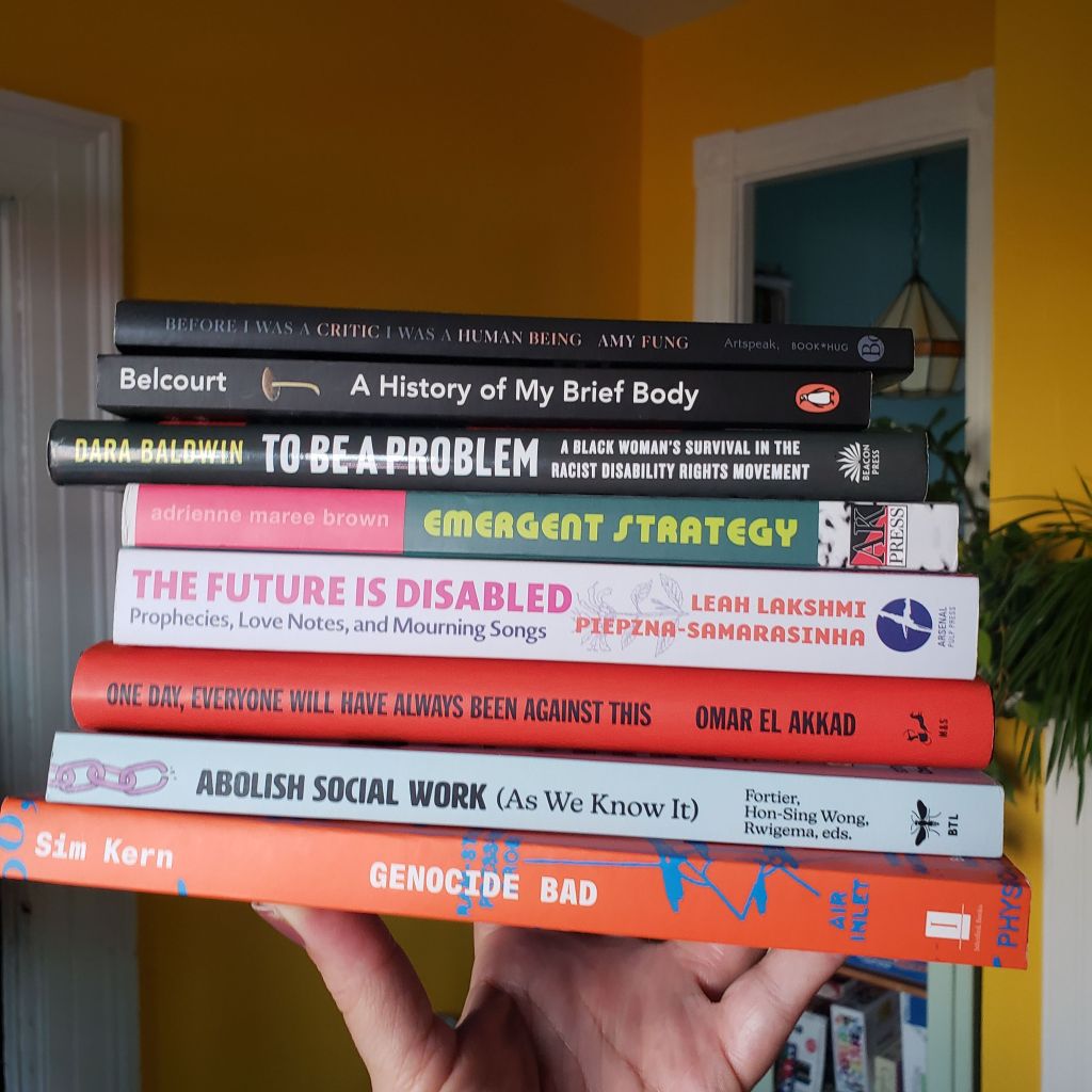 A stack of books is held up by Andreae's hand. In the background, the walls are sunflower yellow, and there are white doorway and window-frame details.  