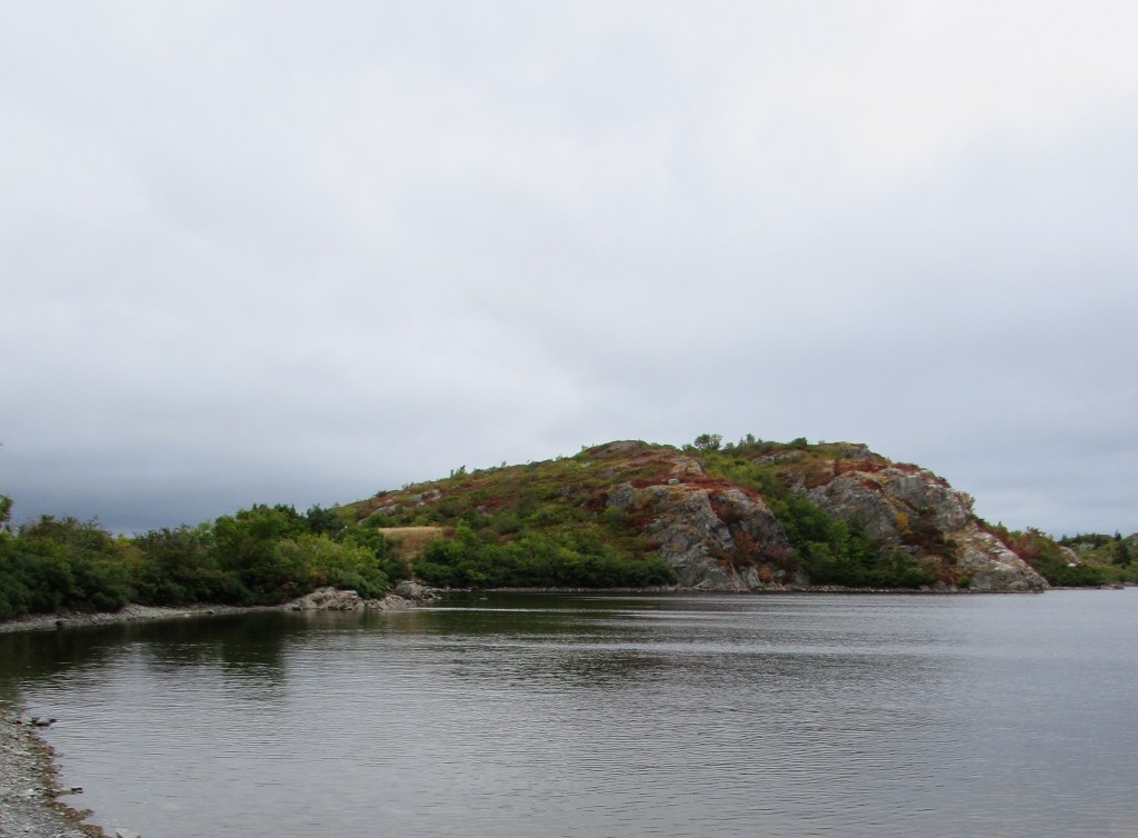 A pond is nestled against a low rocky hill. The hill is covered in low, brushy foliage which is beginning to turn colour for fall. The sky above is cloudy and moody. The water's surface is gently rippled. 