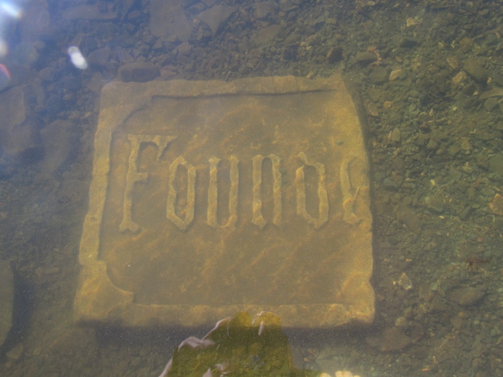 A square slab of flat stone at the bottom of a shallow lake. The image is slightly distorted by the water. The stone looks to have been part of a 19th-century building facade; it reads "Found," but probably said "Founded," followed by a date, before it was broken.