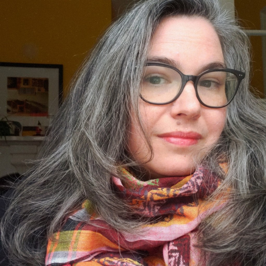 A middle-aged white woman with long greying hair and dark-rimmed glasses looks into the camera. She is indoors and is smiling slightly. She is wearing a patterned scarf in shades of burgundy, pink, mustard, and green. The wall behind her is a shadowy shade of goldenrod.